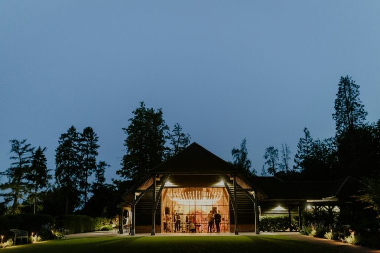 The barn at dusk across the lawn, its glazed gable lit from inside by festoon lights, The Post Barn