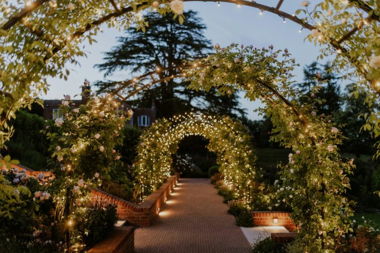 The garden walk of rose arches strung with lights at dusk, The Post Barn