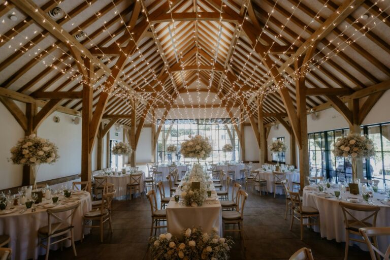 Round tables with white linen and tall flower arrangements under fairy-lit oak trusses, The Post Barn