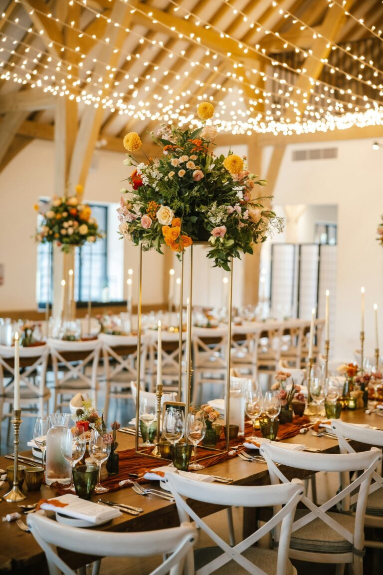 Tall floral centrepieces and candles on long tables beneath a canopy of fairy lights, The Post Barn