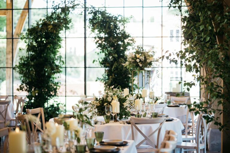 Tables with white linen and candles framed by tall greenery arches against the steel windows, The Post Barn