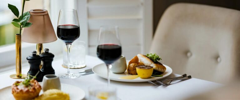 A restaurant table by the window with fish, red wine and a small brass lamp, The Roseate Reading
