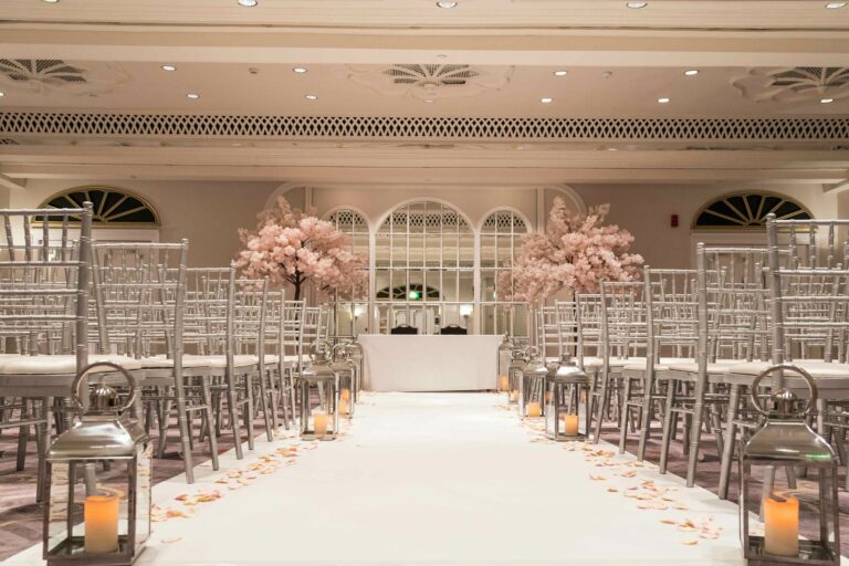 A room set for a ceremony with silver chairs, a white aisle, lanterns and blossom trees either side of the registrar table, The Sheraton Park Lane Hotel