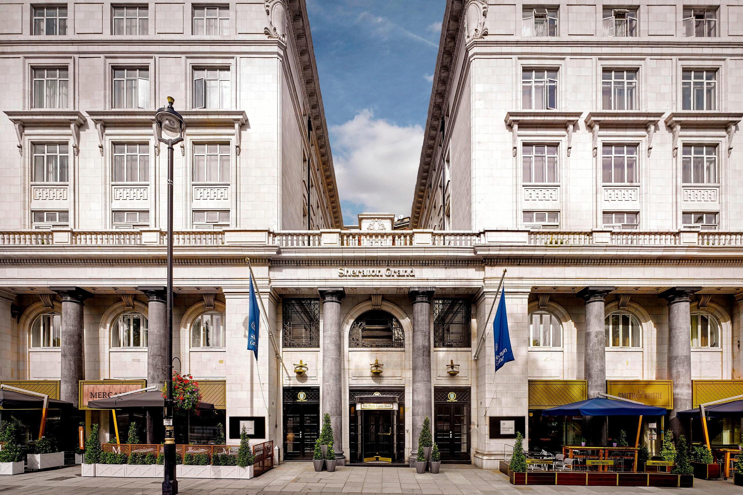The hotel front on Piccadilly, with its pale stone facade, flags over the entrance and terrace seating at street level, The Sheraton Park Lane Hotel