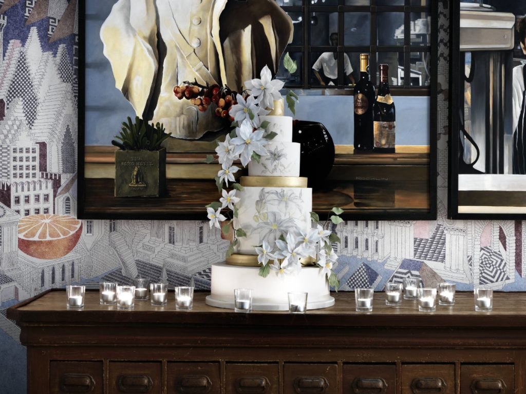 A white tiered wedding cake with sugar flowers on a wooden sideboard against a mural wall