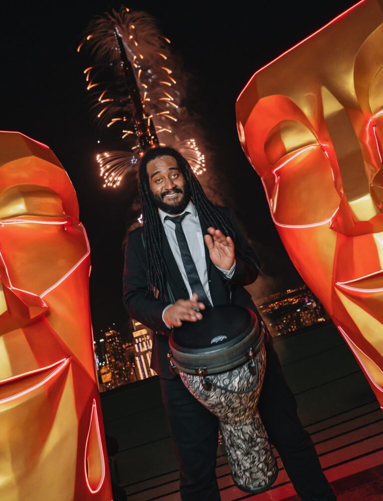 A percussionist with a djembe in front of lit sculptures, The Sway Allstars