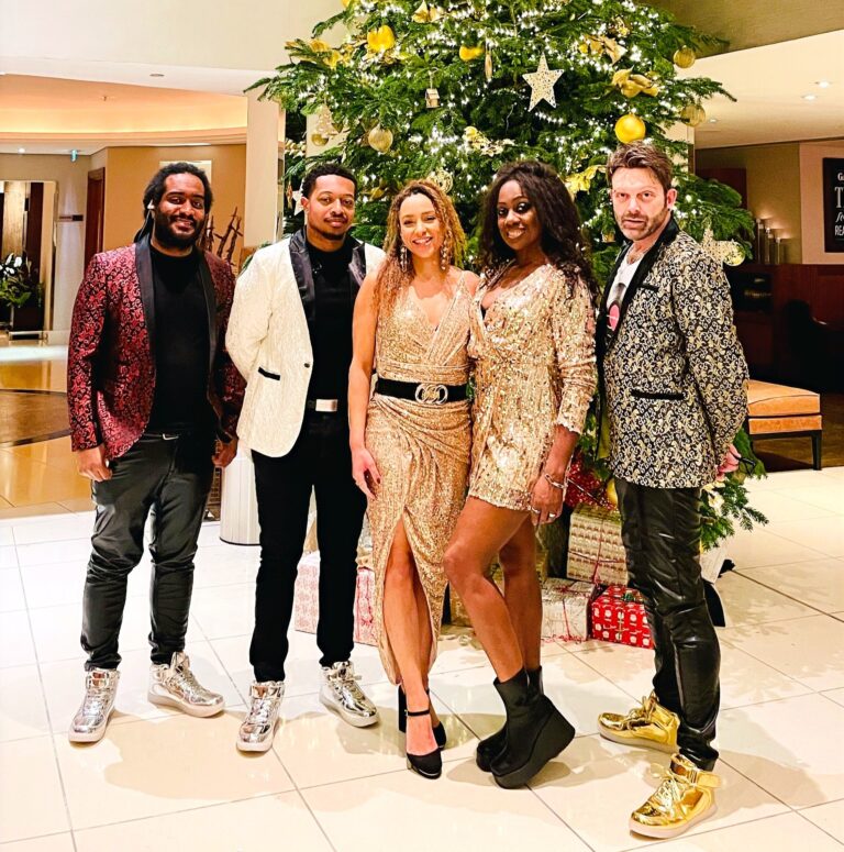 Five band members in sequins and gold in front of a Christmas tree, The Sway Allstars
