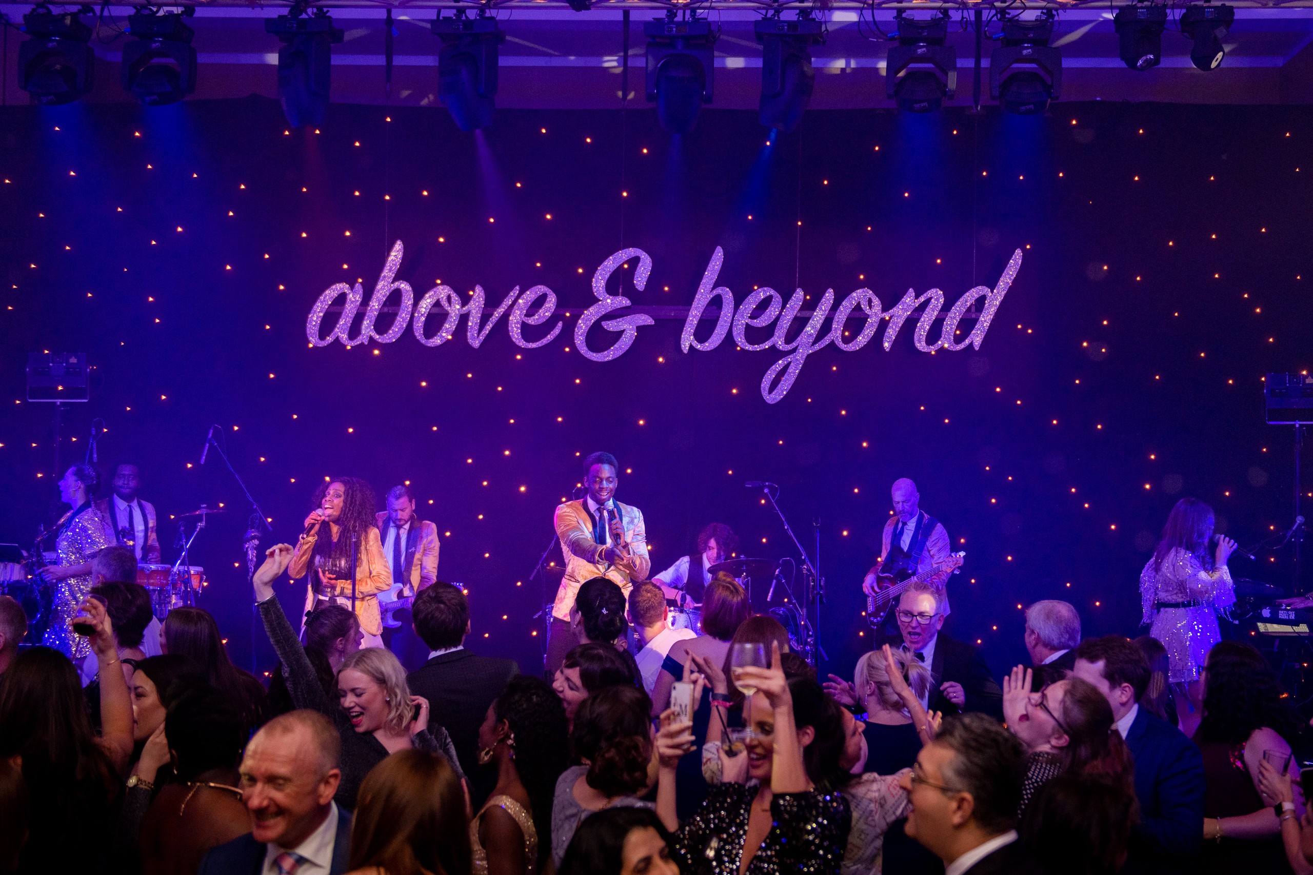 The band on stage at a black tie style party under an Above and Beyond sign, The Sway Allstars