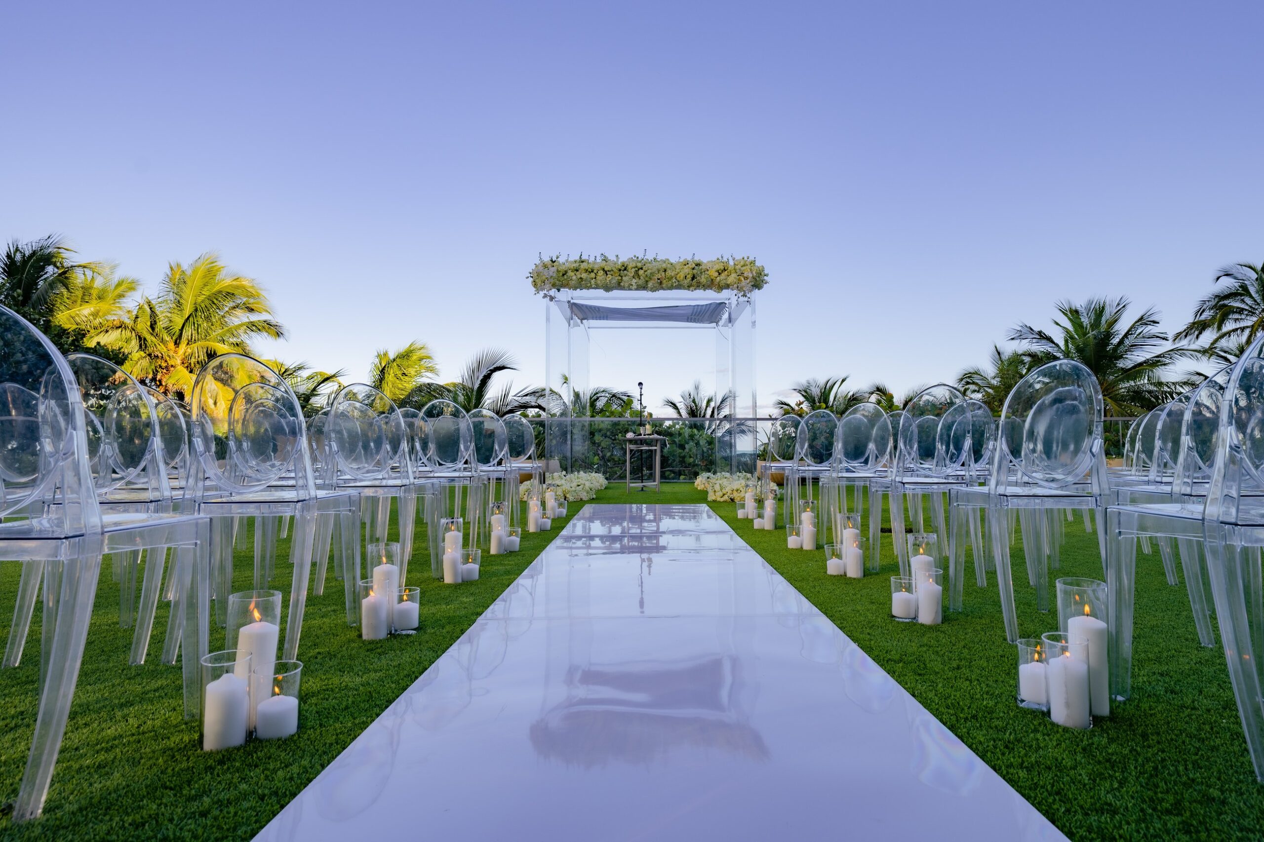 A wedding aisle lined with white chairs on a lawn among palm trees, The Wedding Atelier