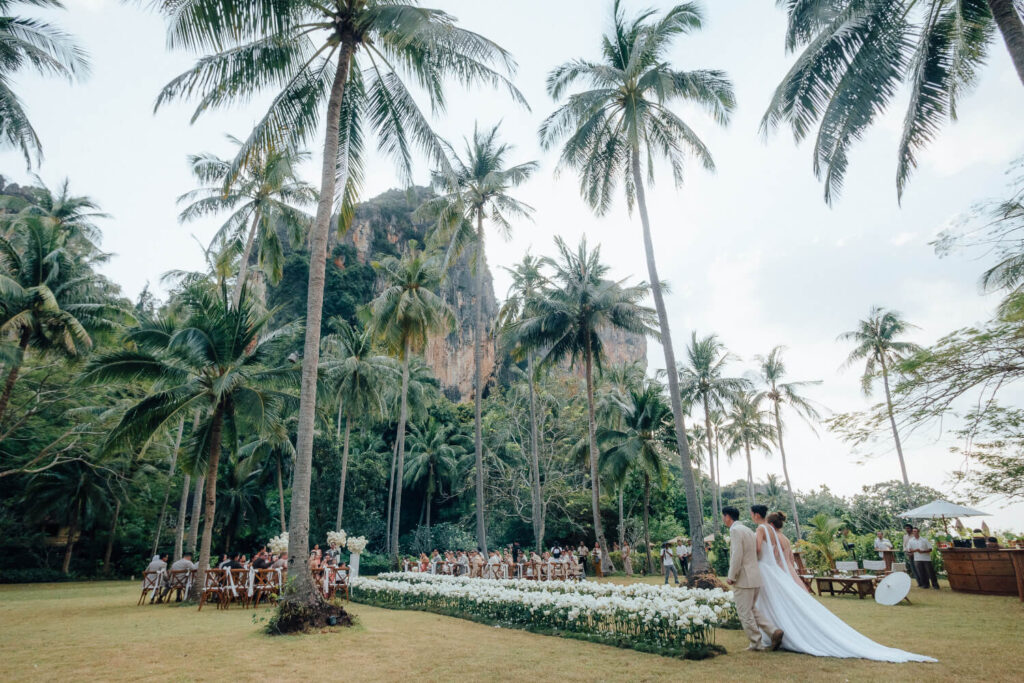 A bride and groom at the end of a white rose aisle among tall palms