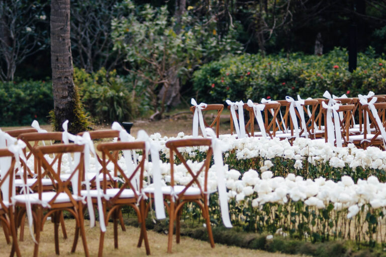 Wooden cross-back chairs with white ribbons beside beds of white flowers, The Wedding Bliss - Thailand
