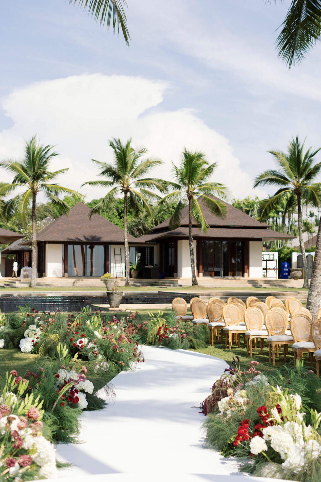 A white aisle edged with red, white and green flowers beside a pavilion and pool, The Wedding Bliss - Thailand