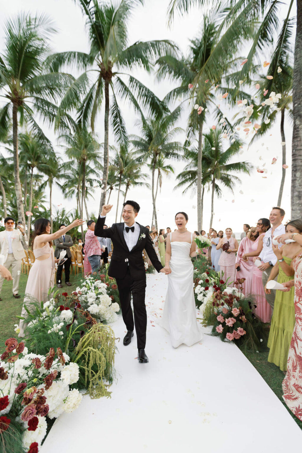 A couple walking the white aisle past guests under the palms