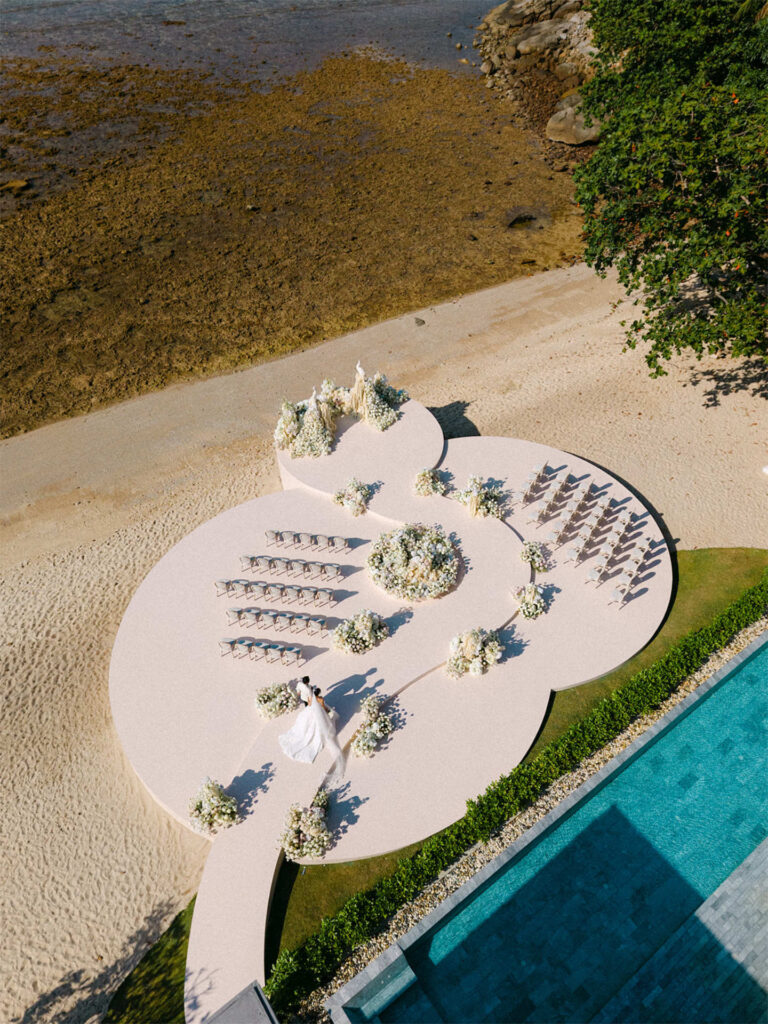 Sculpted ceremony platform with chairs and white flower clusters beside a pool, seen from above, The Wedding Bliss - Thailand