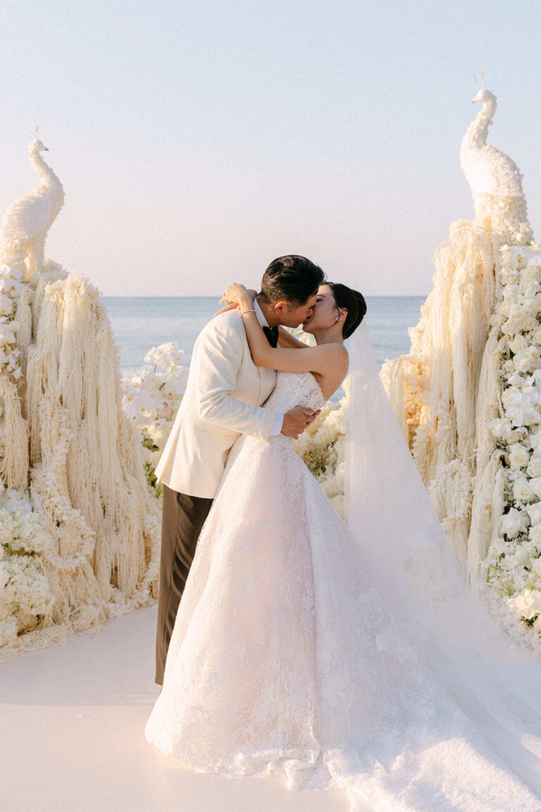 A couple kissing in front of white peacock sculptures and cream candle forms, The Wedding Bliss - Thailand