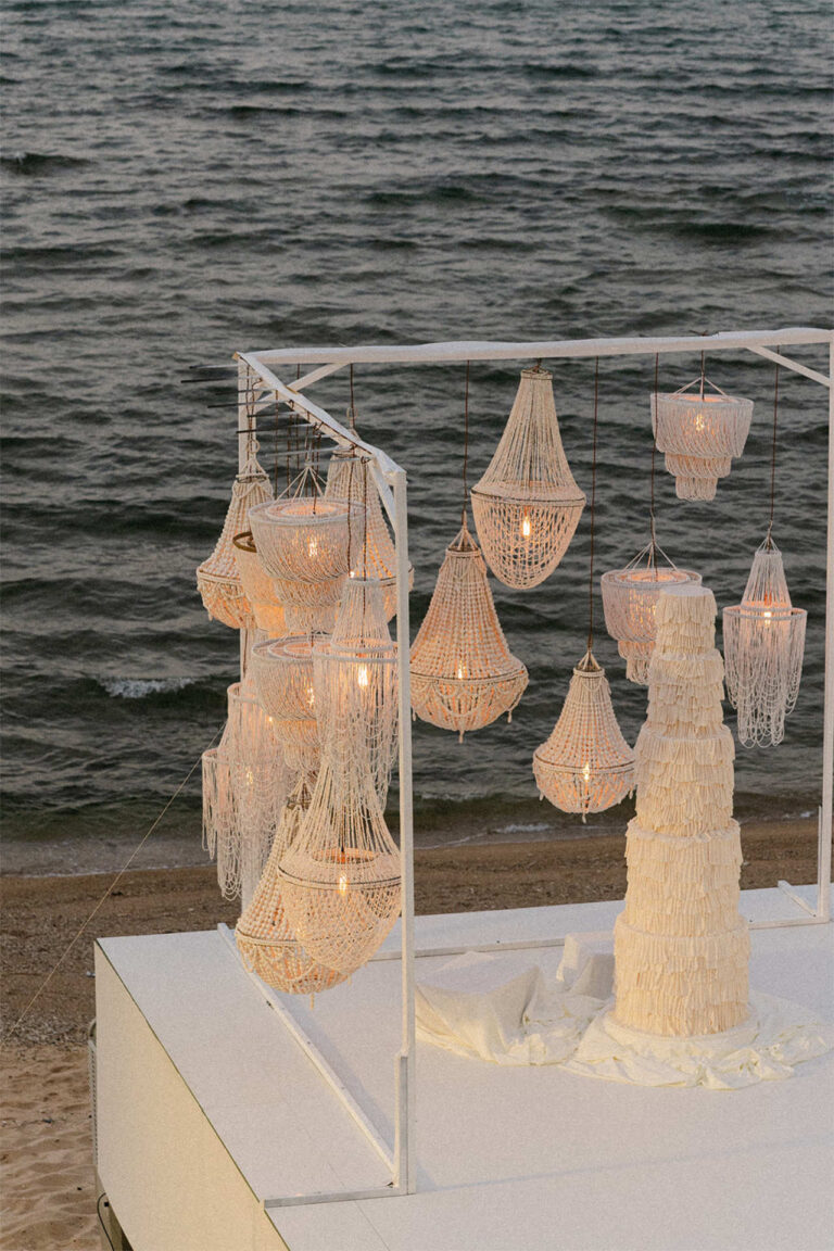 A tiered white cake beneath hanging beaded chandeliers beside the sea, The Wedding Bliss - Thailand