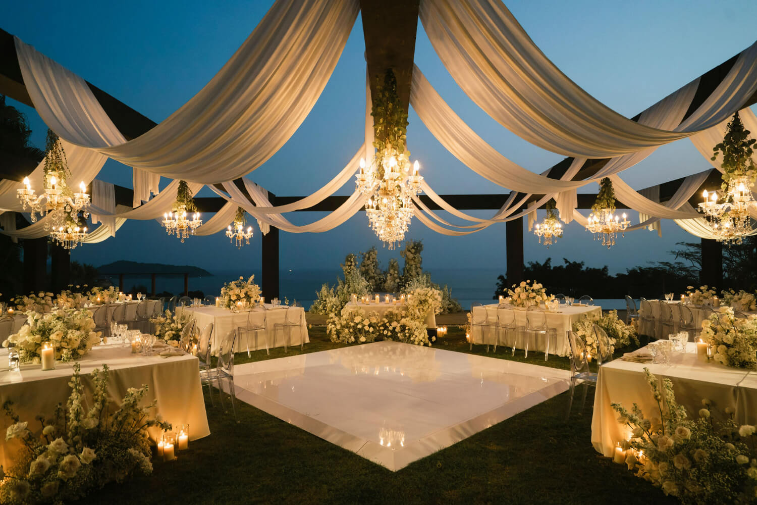 Pavilion draped in white fabric with chandeliers and a lit dance floor at dusk, sea beyond, The Wedding Bliss - Thailand