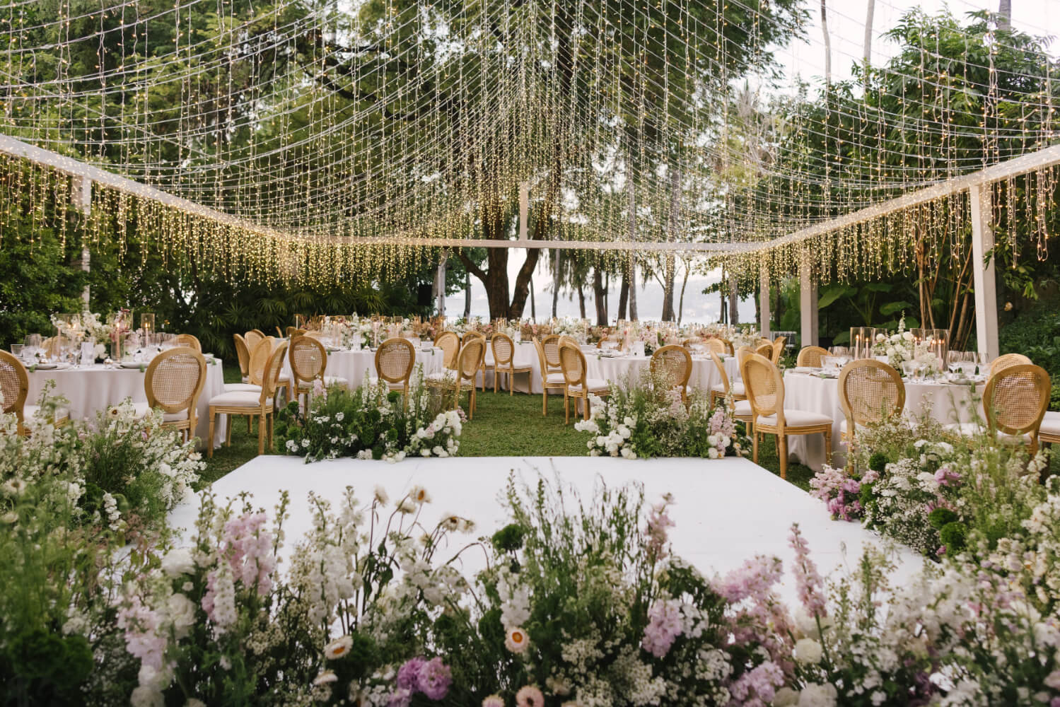 A garden reception under a lattice of hanging lights, with round tables and soft spring flowers, The Wedding Bliss - Thailand