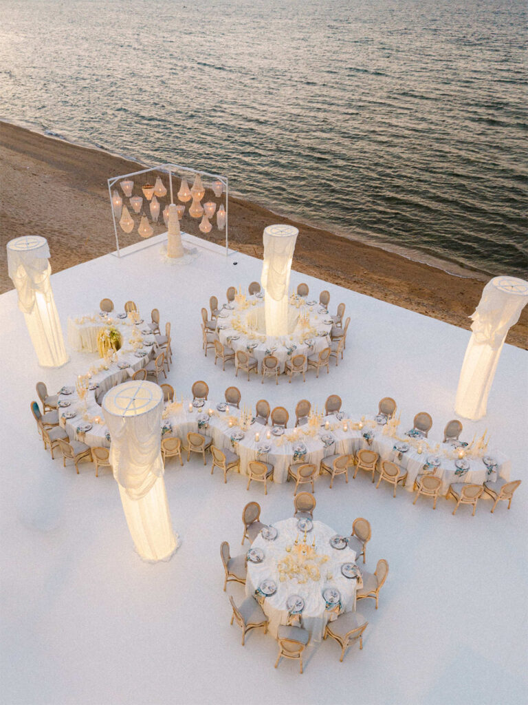 Curved guest tables with glass candle holders on a pale platform by the sea, The Wedding Bliss - Thailand