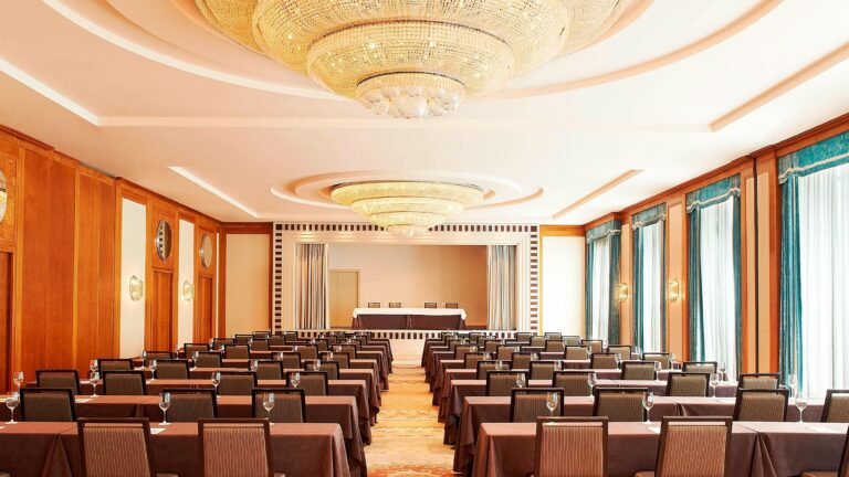 A daylit salon set with rows of chairs beneath twin crystal chandeliers, tall curtained windows, The Westin Valencia