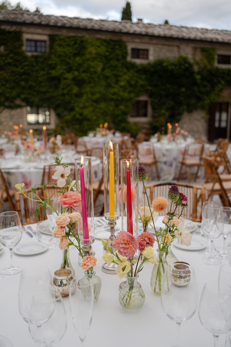 Round tables with coloured candles and loose flowers beside the ivy-covered house at Borgo Pignano, TheKnotInItaly