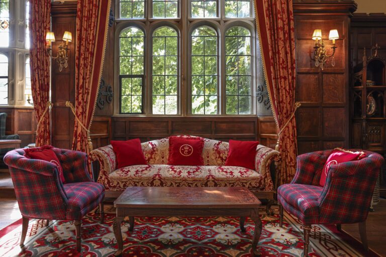 A lounge beneath tall mullioned windows with red sofas and tartan chairs, Thornbury Castle