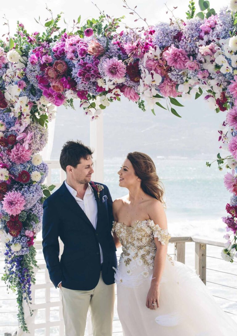 The couple beneath a pink and lilac floral arch on the ceremony deck, Atlantic surf behind, Tintswalo Atlantic