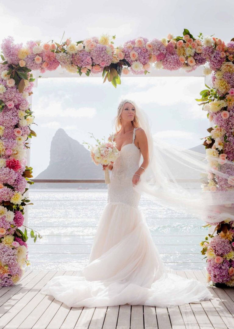 The bride alone beneath the floral arch on the pool deck, the Sentinel framed through it, Tintswalo Atlantic