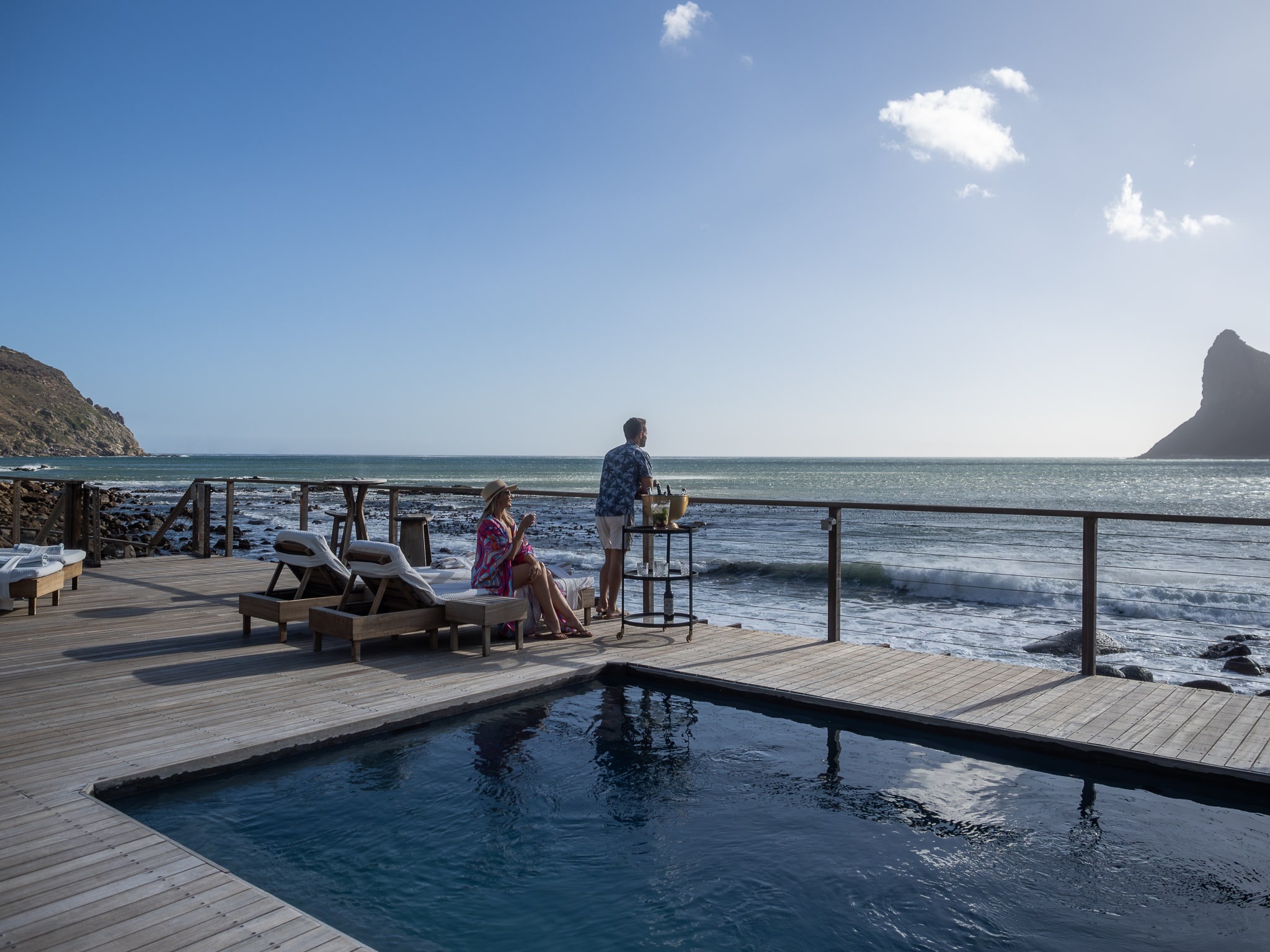 The pool deck at the water's edge, guests at the rail, the Sentinel across the bay, Tintswalo Atlantic