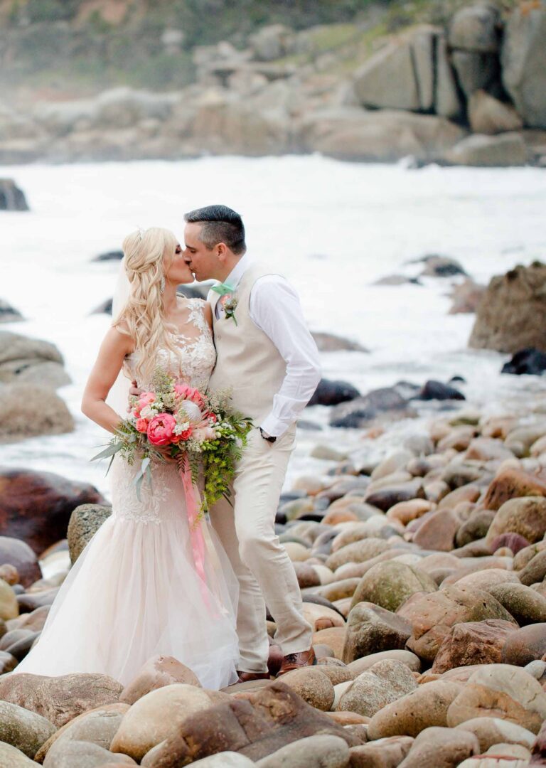 The couple kissing on the boulder beach, the bride carrying protea and roses, Tintswalo Atlantic