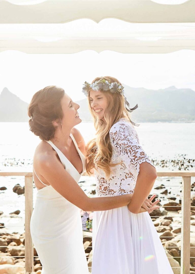 Two brides embracing on the deck rail above the pebbled shore, Tintswalo Atlantic