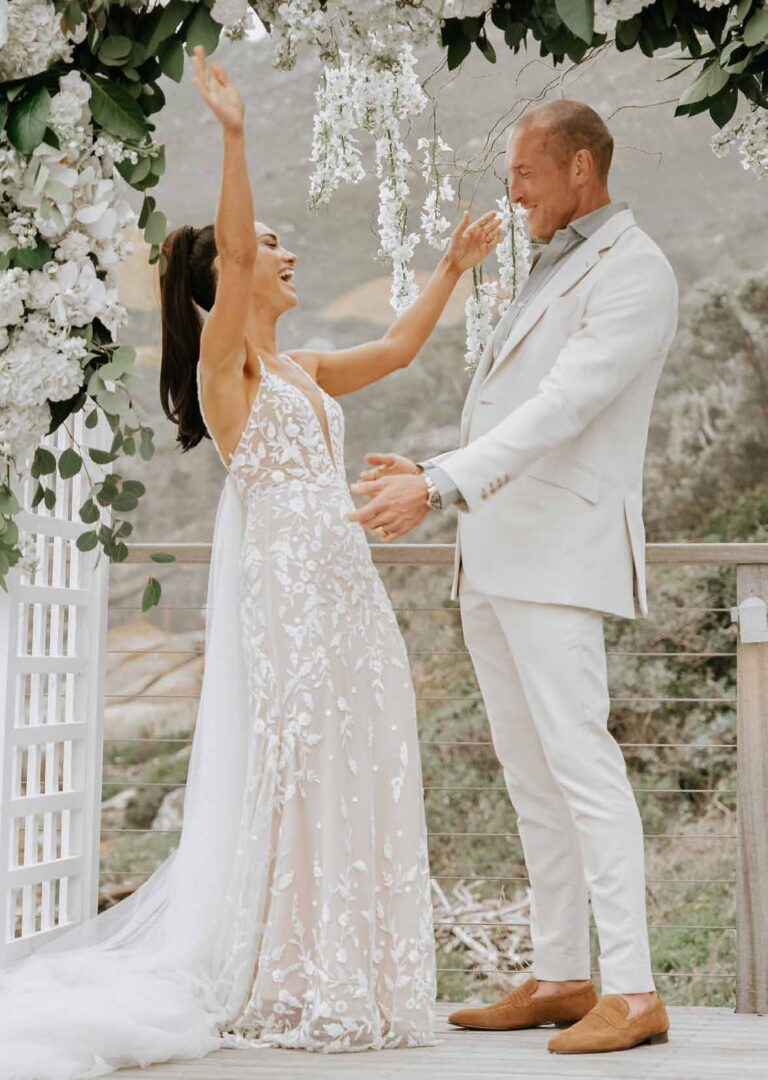 The couple beneath a hanging floral installation, the bride reaching up, Tintswalo Atlantic