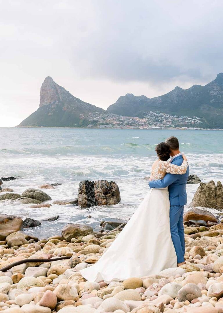 The couple embracing on the boulder beach, the Sentinel rising behind, Tintswalo Atlantic