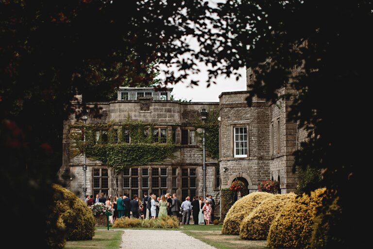 The hall's stone facade across the lawn, guests gathered, Tissington Hall