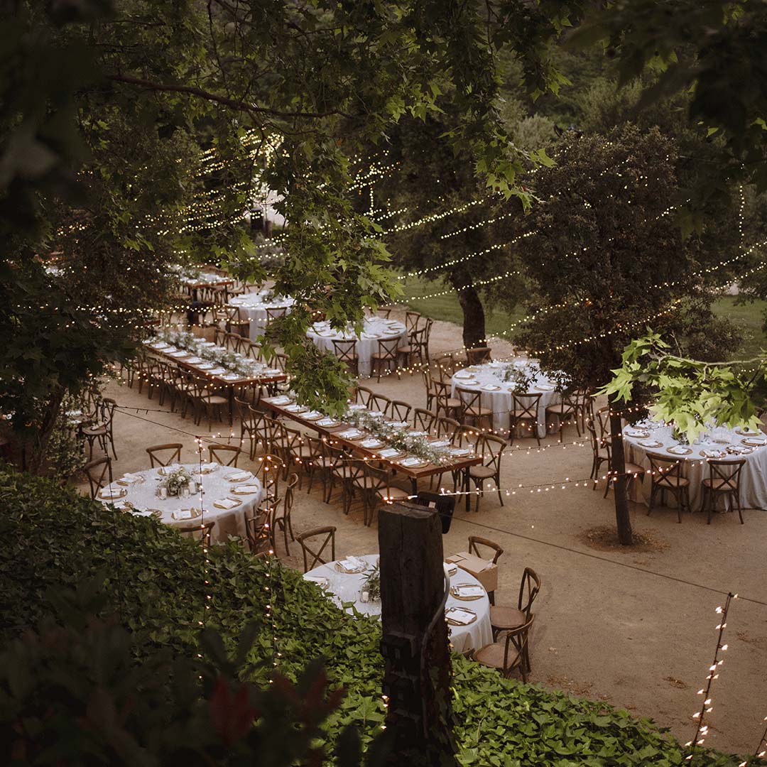 Long and round dinner tables laid among the trees under string lights, seen from above, Torre Sever
