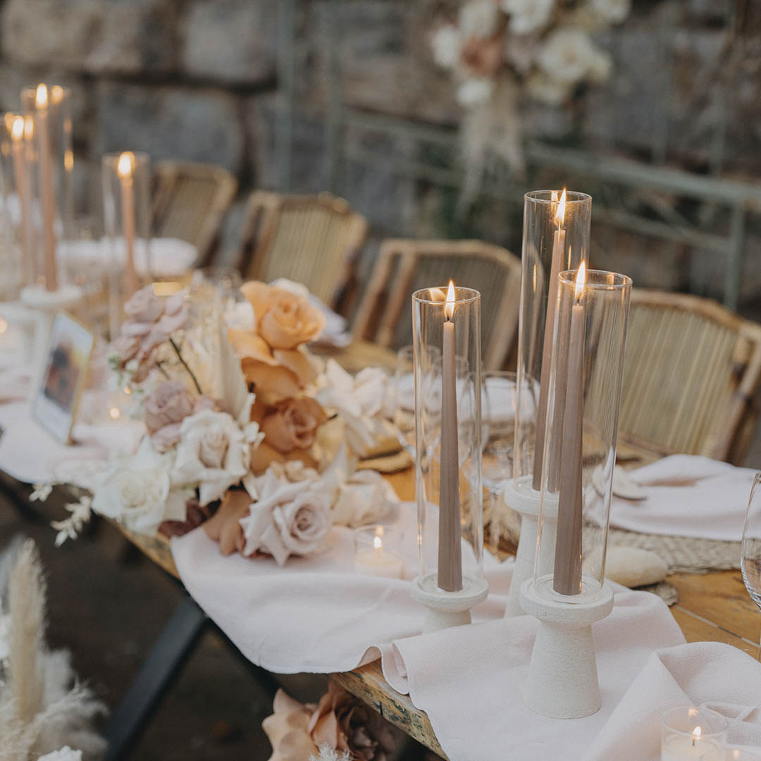 A dinner table with pillar candles and cream roses, with a stone wall behind, Torre Sever