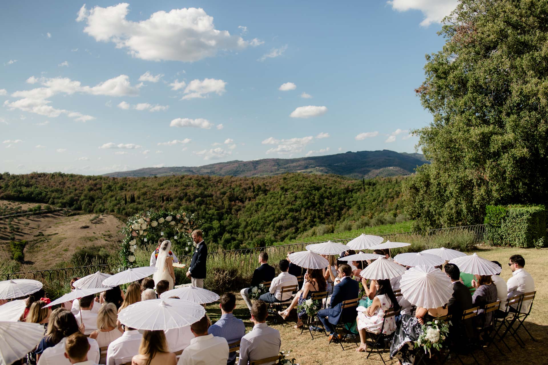 Guests under white paper parasols at an outdoor ceremony, the couple at a flower arch with the Tuscan hills behind, Tuscan DMC