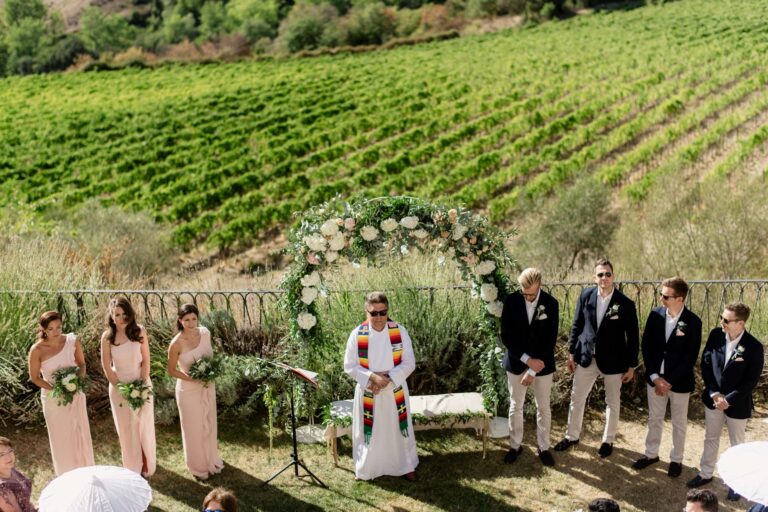 Ceremony at a flower arch above a vineyard, bridesmaids and groomsmen either side of the officiant, Tuscan DMC