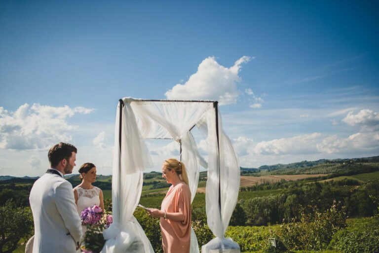 An outdoor ceremony under white drapes, the officiant reading to the couple, hills beyond, Tuscan DMC