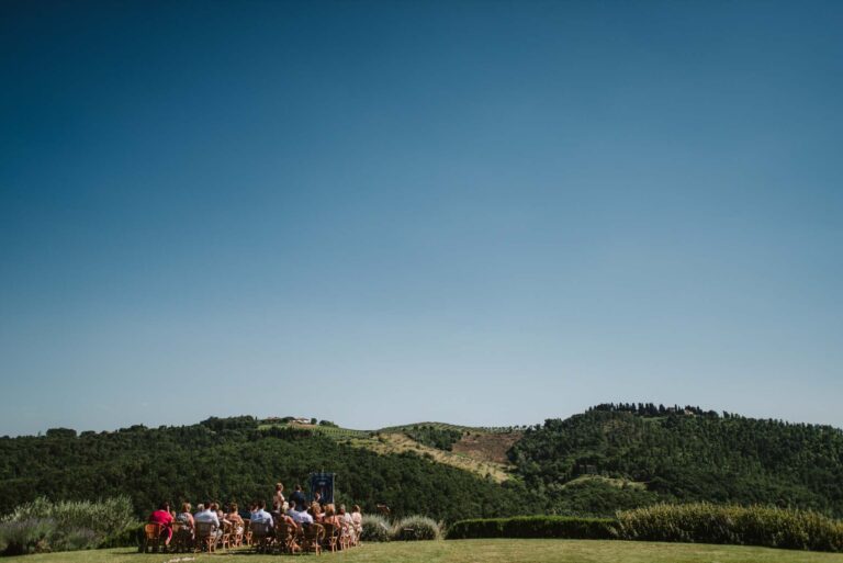 A small outdoor ceremony on a lawn, seen wide against wooded green hills, Tuscan DMC