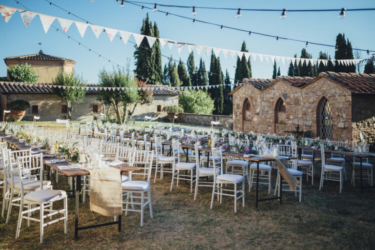 Long dinner tables laid in a garden under bunting and festoon lights, cypresses behind, Tuscan DMC