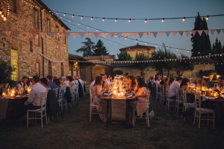 Guests at candlelit dinner tables at dusk beside a stone building, bunting and lights overhead, Tuscan DMC