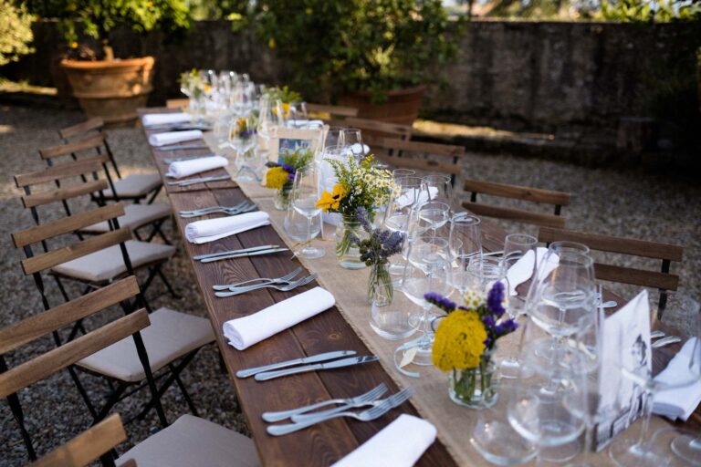 A long wooden table on a gravel terrace, wildflowers in small bottles and folding chairs, Tuscan DMC