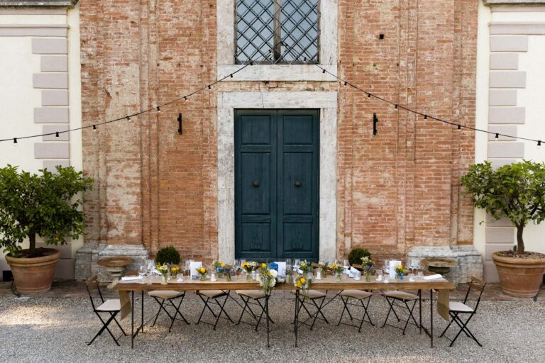 A dinner table laid in front of a brick villa facade, lemon trees in pots either side, Tuscan DMC