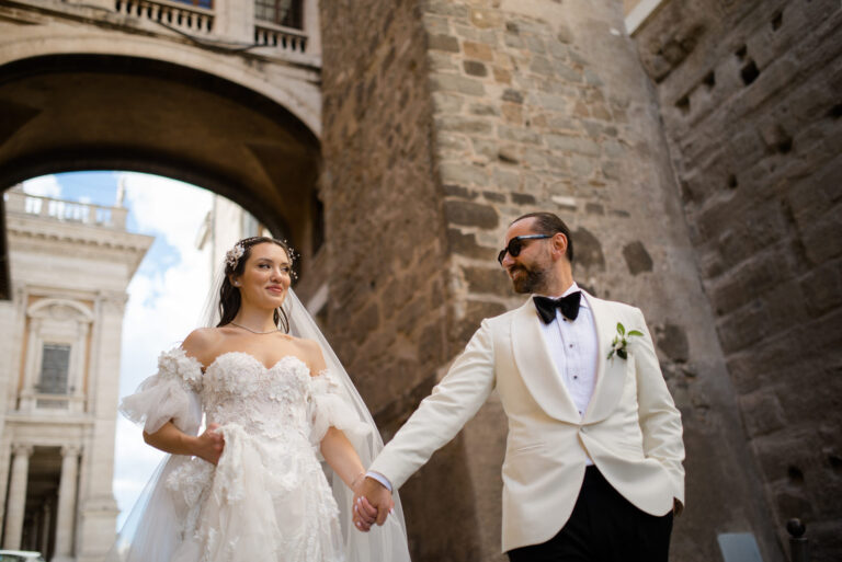 Couple holding hands beneath a stone arch in Rome, Vera Weddings