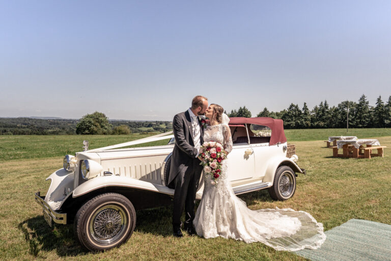 A kiss beside a vintage wedding car in a summer meadow, England, Victoria Amrose Photography
