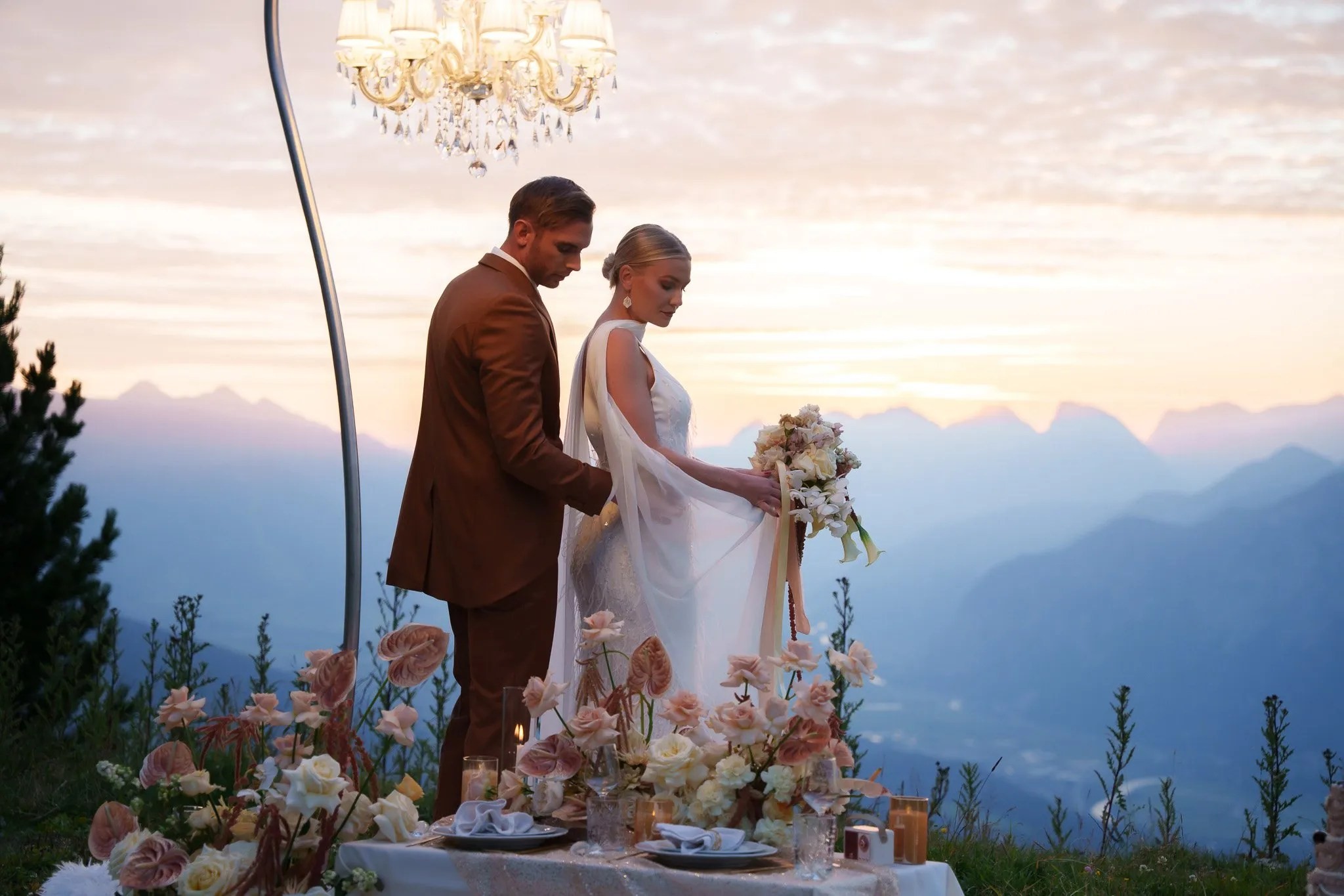Couple at a mountain-top elopement table with a hanging chandelier and soft alpine light, Vienna Picnics