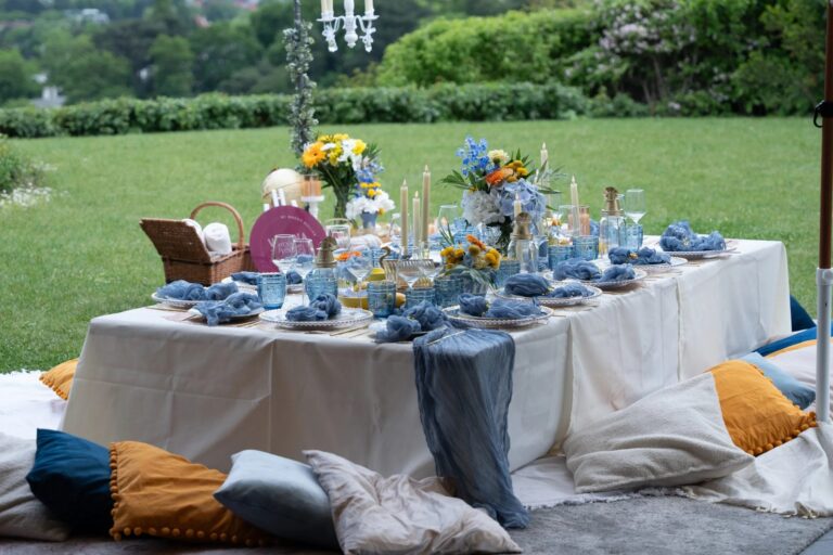 Styled picnic table with blue glassware, flowers and cushions on the lawn, Vienna Picnics