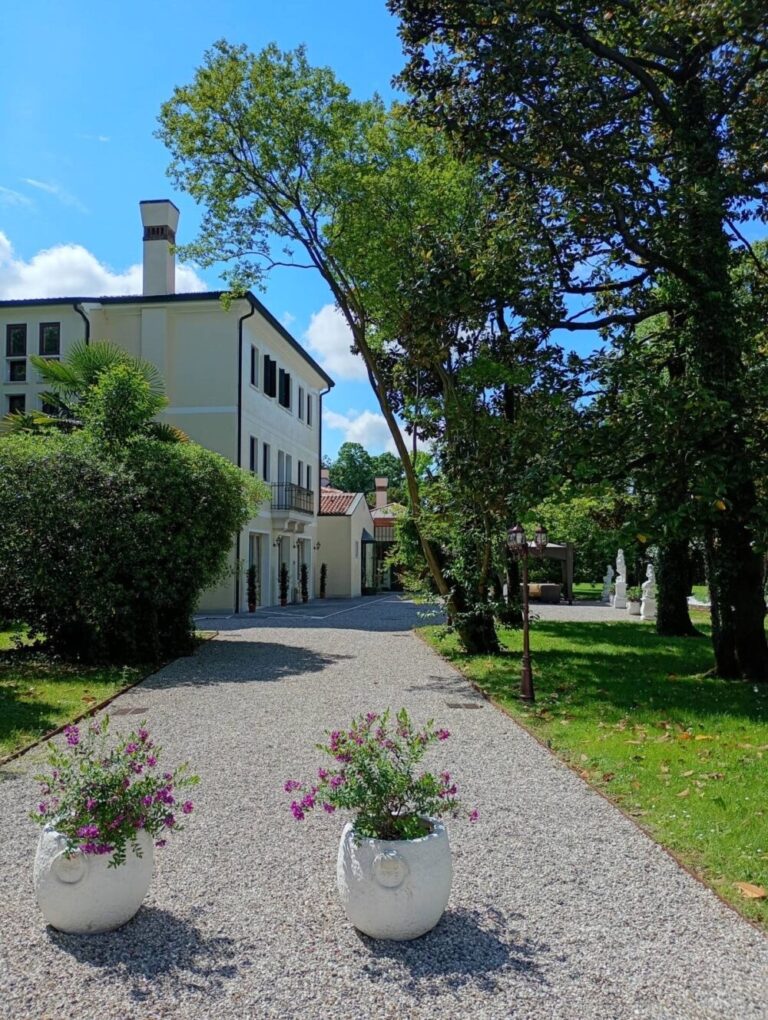 The gravel driveway to the villa, potted flowers and trees leading to the pale facade, Villa Pace Park Hotel Bolognese