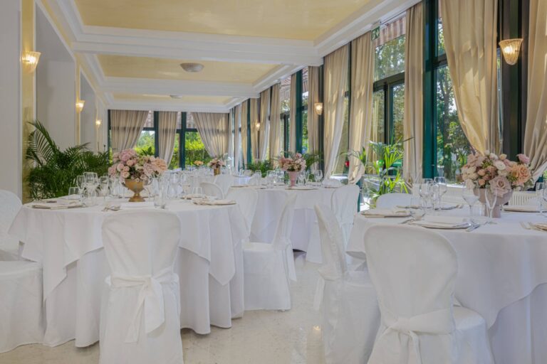 A veranda laid for a wedding, pastel florals and dressed chairs beneath a soft golden ceiling, the park beyond the glass, Villa Pace Park Hotel Bolognese